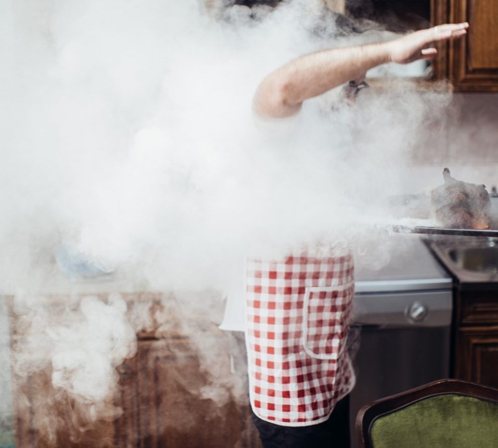 A man in an apron cooking in the kitchen near the oven full of smoke. Bertolli Extra Virgin Olive Oil. Best Olive Oil Brands.