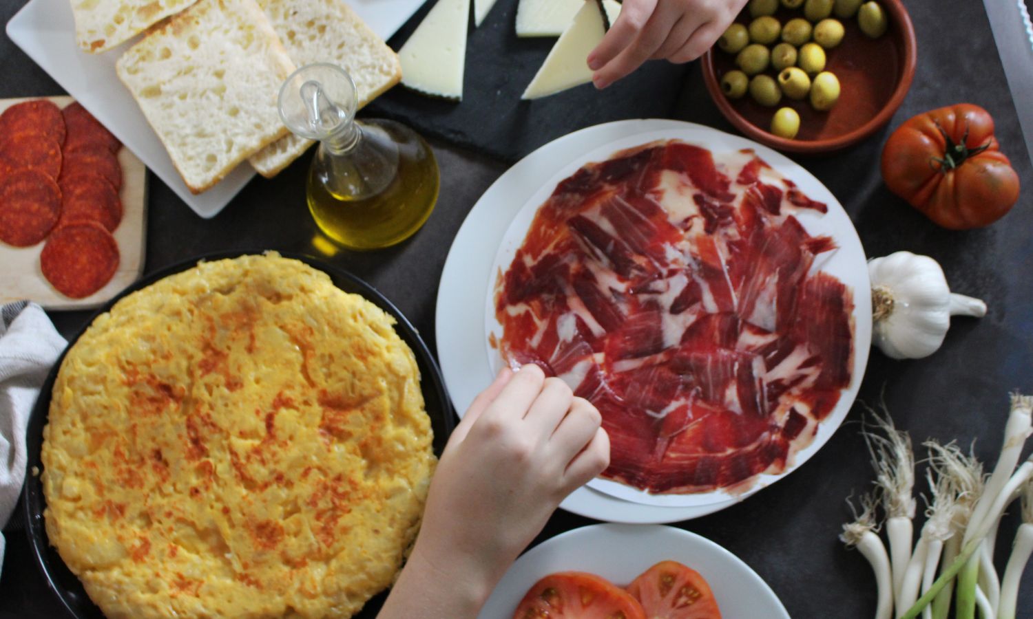 Kitchen table with food, namely jar of olive oil, olives, Bertolli extra virgin olive oil, tomatoes, garlic, bread and cheese